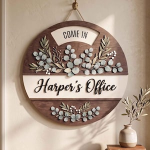 Könnte beinhalten: Ein rundes Wandschild aus Holz mit dunkelbraunem Finish und einem cremefarbenen horizontalen Mittelstreifen. Der Text "COME IN" steht oben, "Harper's Office" ist in schwarzer Schreibschrift gehalten. Das Schild ist mit hellgrünen Eukalyptusblättern verziert.