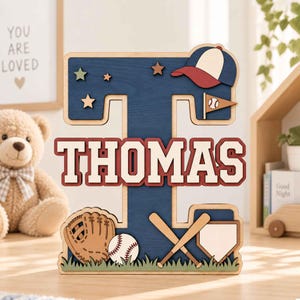 Könnte beinhalten: Ein großer hölzerner Buchstabe T mit dunkelblauer Oberfläche und dem Namen THOMAS in weißen und roten Blockbuchstaben. Das Schild ist mit Baseball-Motiven verziert, darunter eine Kappe, ein Handschuh, ein Ball, ein Schläger und eine Homeplate.