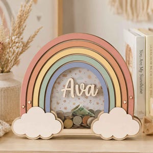 Puede incluir: Una hucha de madera con forma de arcoíris y panel frontal transparente. El arcoíris presenta arcos en tonos pastel rosa, amarillo, verde y azul. El nombre Ava está escrito en letras blancas en el centro. La base tiene dos nubes blancas.