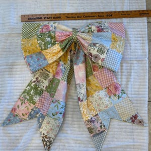 May include: A large fabric bow made of patchwork squares featuring various floral, gingham, and butterfly patterns in pastel pink, yellow, blue, and green. A wooden ruler measuring 32 inches is placed above the bow for scale.