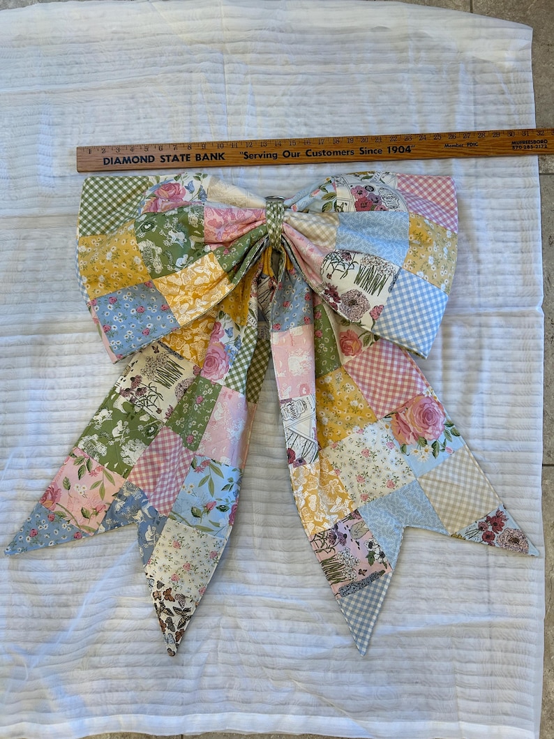May include: A large fabric bow made of patchwork squares featuring various floral, gingham, and butterfly patterns in pastel pink, yellow, blue, and green. A wooden ruler measuring 32 inches is placed above the bow for scale.