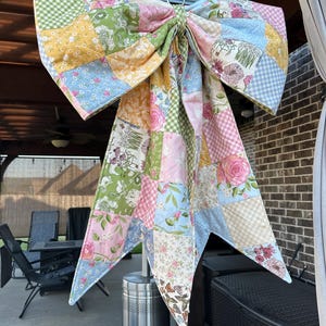 May include: A large decorative bow constructed from a patchwork of quilted fabric squares. The fabric features a variety of floral, gingham, and butterfly patterns in soft pastel shades of pink, blue, yellow, green, and white.