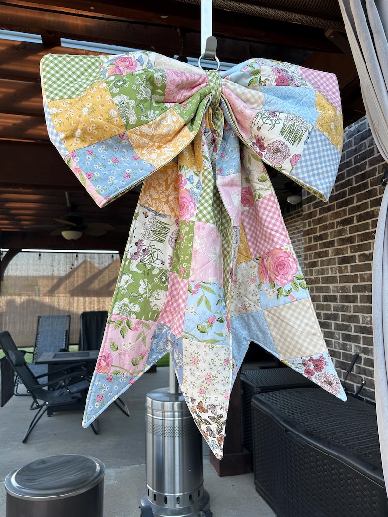 May include: A large decorative bow constructed from a patchwork of quilted fabric squares. The fabric features a variety of floral, gingham, and butterfly patterns in soft pastel shades of pink, blue, yellow, green, and white.