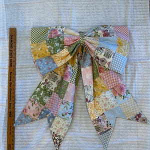 May include: A large fabric bow made of patchwork squares featuring floral, gingham, and solid patterns in pink, yellow, blue, and green. A wooden ruler placed next to the bow shows it measures approximately 12 inches in height.