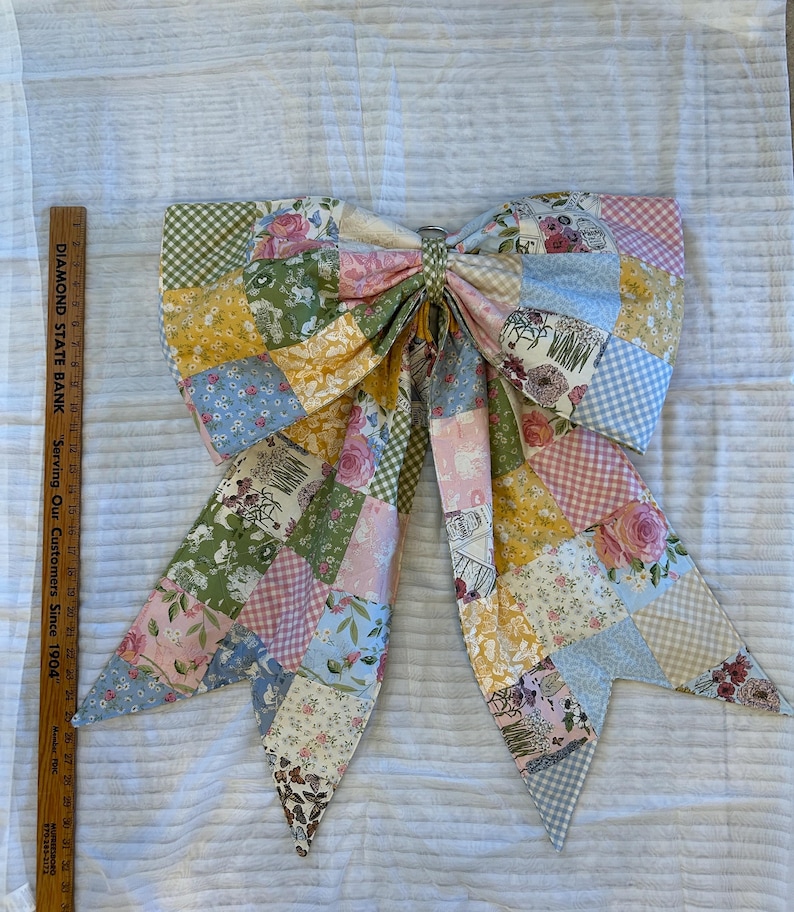 May include: A large fabric bow made of patchwork squares featuring floral, gingham, and solid patterns in pink, yellow, blue, and green. A wooden ruler placed next to the bow shows it measures approximately 12 inches in height.