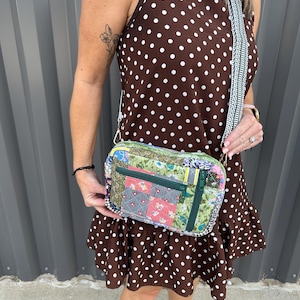 May include: A rectangular crossbody bag featuring a patchwork design with various floral and geometric fabric patterns in shades of green, red, and blue. The bag includes a dark green zippered front pocket and a wide, woven, patterned shoulder strap.