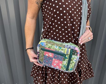 May include: A rectangular crossbody bag featuring a patchwork design with various floral and geometric fabric patterns in shades of green, red, and blue. The bag includes a dark green zippered front pocket and a wide, woven, patterned shoulder strap.