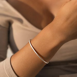 Pode incluir: Uma pulseira fina de tom prateado com uma superfície texturizada e ranhurada com linhas verticais repetitivas. A banda metálica tem um acabamento polido que reflete a luz, criando um contraste entre as cristas elevadas e as áreas rebaixadas.