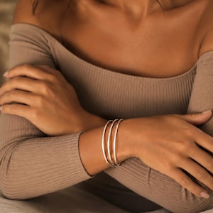 Può includere: Un set di tre bracciali rigidi sottili in metallo color argento lucido indossati al polso. I bracciali presentano un profilo liscio e arrotondato con una finitura metallica riflettente, creando un aspetto sovrapposto sul braccio.
