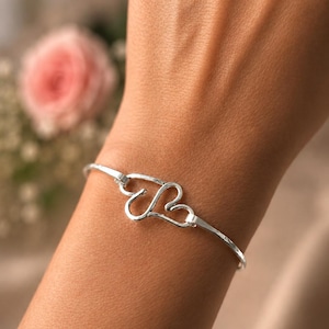 Pode incluir: Uma pulseira de tom prateado com um design central de dois contornos em forma de coração entrelaçados. A fina banda de metal polido conecta-se ao motivo de coração em ambos os lados, criando uma forma circular contínua no pulso.