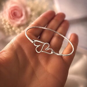 May include: A silver-toned bangle bracelet featuring two interlocking heart-shaped charms at the centre. The metal band has a textured, hammered finish, and the two hearts are connected to the circular wire frame, creating a continuous loop design.