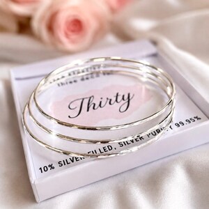 Könnte beinhalten: Drei silberfarbene, gehämmerte Armreifen auf einer weißen Geschenkbox. Die Box zeigt den Schriftzug Thirty in einer Schreibschrift sowie den aufgedruckten Text 10% SILVER FILLED, SILVER PURITY 99.95% und weiteren teilweise sichtbaren Text.