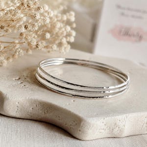 Pode incluir: Um conjunto de três pulseiras finas de cor prateada com um acabamento martelado e texturizado. As pulseiras circulares estão empilhadas, apresentando uma superfície metálica reflexiva que capta a luz de forma uniforme.