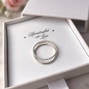 Op de afbeelding: Twee zilveren ringen in een witte vierkante geschenkdoos. De ene ring heeft een gladde, gepolijste band, terwijl de andere een gedraaide, touwachtige textuur heeft. De binnenkant van de doos is bedrukt met de tekst Handcrafted with Love.