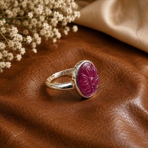 Op de afbeelding: Een zilveren ring met een gepolijste, ovaalvormige, diepmagenta edelsteen die is uitgesneden met een bloemmotief. De ringband is glad en metaalachtig en houdt de edelsteen vast in een eenvoudige zetting. De ring is scherp in beeld gebracht.