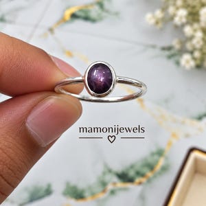 Könnte beinhalten: Ein Silberring mit einem polierten, ovalen, tiefvioletten Stern-Edelstein in einer glatten Fassung. Das Ringband ist dünn und metallisch. Der Text mamonijewels mit einem kleinen Herzsymbol ist unter dem Ring im Bild zu sehen.