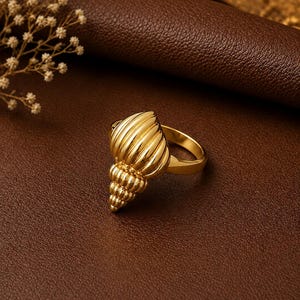 Puede incluir: Un anillo de tono dorado que presenta un diseño de concha marina tridimensional prominente. La concha tiene una superficie pulida y reflectante con crestas verticales y una punta espiralada y cónica. El aro es liso, sólido y circular.