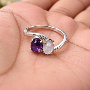 May include: A silver bypass ring featuring two oval-shaped cabochon gemstones. One stone is a deep purple amethyst, and the other is a translucent white moonstone. The stones are set in polished silver prongs on a smooth, slender silver band.