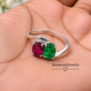 May include: A silver ring featuring two oval-cut gemstones, one deep red and one vibrant green, set side-by-side with small clear accent stones. The band has a bypass design. The text MamoniJewels with a heart symbol is printed on the image.