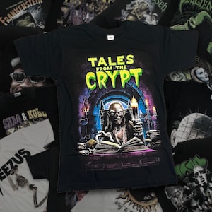 Op de afbeelding: Een zwart t-shirt met korte mouwen en een grafische print van de Crypt Keeper uit Tales from the Crypt. Het ontwerp toont het personage dat een kaars boven een open boek in een stenen boog houdt, met de tekst Tales from the Crypt in het groen.