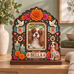 May include: A decorative wooden picture frame featuring a central photo of a brown and white spaniel. The frame is adorned with colorful marigolds, two white doves, paw prints, hearts, two lit teal candles, and a figurine of a spaniel with the name BELLA.