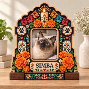May include: A decorative wooden picture frame featuring a central photo of a Siamese cat. The frame is adorned with colorful marigold flowers, papel picado banners, paw prints, and a candle. The name SIMBA is printed on a plaque at the bottom.