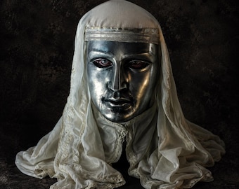 May include: A metallic silver face mask with human-like features, including defined eyes, a nose, and lips. The mask is draped in a sheer, off-white fabric head covering that features a decorative band across the forehead and lace trim edges.