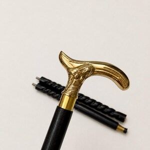 Op de afbeelding: Een wandelstok met een gepolijst goudkleurig metalen handvat in de vorm van een gestileerd figuur. Het handvat is bevestigd aan een gladde, donkerzwarte houten schacht. De stok is ontworpen in twee afneembare delen met een zichtbaar metalen verbindingsstuk.