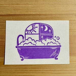 May include: A purple ink block print on white paper featuring a clawfoot bathtub filled with bubbles. Above the tub is a window divided into four panes, showing a sun on the left and a crescent moon with stars on the right side of the frame.