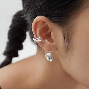 Puede incluir: Un ear cuff de plata con una forma lisa, redondeada y ligeramente irregular colocado en el cartílago superior, junto con un pendiente de aro de plata a juego que presenta un dije de metal pulido en forma de gota.
