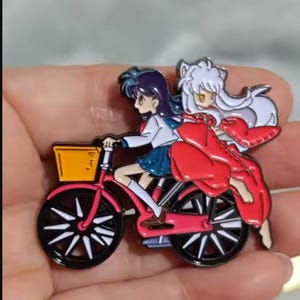 May include: A metal enamel pin featuring two anime-style characters riding a pink bicycle. One character has long dark hair and a blue skirt, while the other has white hair, animal ears, and a flowing red garment. The bicycle has a yellow basket.