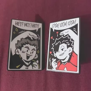 Peut inclure: Deux broderies émaillées rectangulaires représentant des personnages de style anime. La broche gauche montre un personnage en maillot noir et jaune avec le texte HEY! HEY! HEY! La droite montre un personnage en rouge avec OYA! OYA! OYA! et des cœurs.