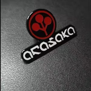 May include: A black and red enamel pin featuring a circular logo with three red spheres on a stem above the text ARASAKA in a stylized white font. The pin has a polished finish and a distinct geometric design with clean lines and high contrast.