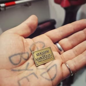 Może przedstawiać: Mała prostokątna metalowa tabliczka w kolorze mosiądzu z wytłoczonym napisem "NELSON AND MURDOCK ATTORNEYS AT LAW" oraz czterema małymi okrągłymi nitami w każdym rogu, prezentowana jako płaska, polerowana replika tabliczki biurowej.