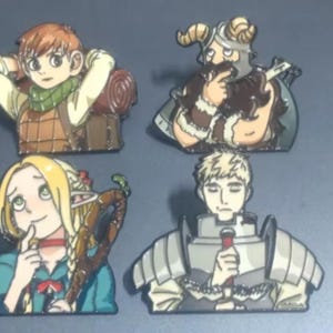 May include: Four enamel pins featuring anime-style characters. Top left: a person with orange hair and a green scarf. Top right: a bearded figure with horns. Bottom left: a blonde elf with a wooden staff. Bottom right: a person in silver armour.