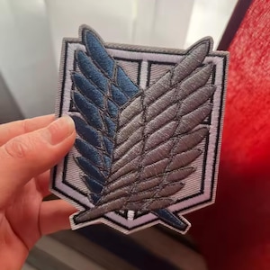 May include: A shield-shaped embroidered patch featuring the Wings of Freedom emblem. The design displays two overlapping wings, one in dark blue and one in grey, set against a white background with a thick grey border and black outlines.