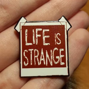 Peut inclure: Une broche en émail rectangulaire en forme de photographie Polaroid avec un centre rouge foncé et une bordure blanche. Le texte "LIFE IS STRANGE" est imprimé en lettres capitales blanches et usées au centre de la zone carrée rouge.