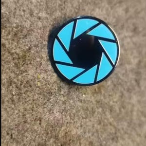 Op de afbeelding: Een ronde emaille speld met een zwart midden, omringd door acht helderblauwe driehoekige segmenten in een camera-diafragmapatroon, omsloten door een dunne zwarte metalen rand die de cirkelvormige omtrek van de speld markeert.