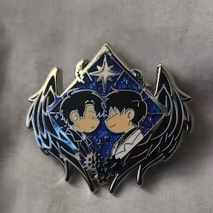 Peut inclure: Une broche en émail métallique représentant deux personnages stylisés aux cheveux sombres se faisant face sur un fond bleu pailleté. Le motif est encadré par des ailes noires et une étoile à quatre branches, avec des détails en forme d'engrenage.