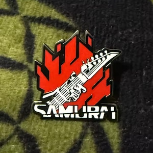 Könnte beinhalten: Ein Metall-Emaille-Pin mit einem stilisierten roten Flammendesign hinter einer weißen und schwarzen E-Gitarren-Kopfplatte. Das Wort SAMURAI ist in fetten, weißen Blockbuchstaben am unteren Rand des Pin-Designs aufgedruckt.