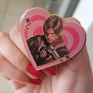 May include: A heart-shaped enamel pin featuring a central illustration of a character with brown hair wearing a brown jacket with a fur collar, holding a silver handgun. The background of the pin consists of concentric pink and light pink heart rings.