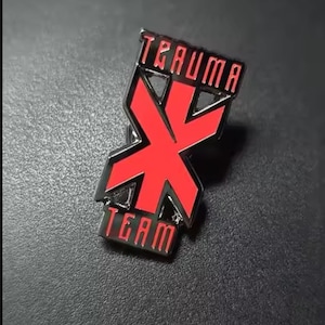 May include: A black and red metal enamel pin featuring a stylized X symbol in the center. The word TRAUMA is printed in red capital letters above the X, and the word TEAM is printed in red capital letters below the X, all set against a black background.