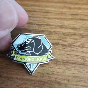 May include: A metal enamel pin shaped like a diamond with a light blue and white geometric background. A black silhouette of a dog head is centered on the pin. A yellow banner across the bottom features the white text DIAMOND DOGS.