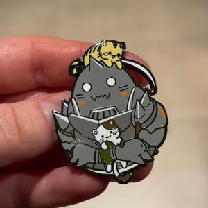Pode incluir: Um broche de esmalte metálico apresentando uma figura de armadura cinza com uma expressão de surpresa, um pequeno gato amarelo listrado empoleirado na cabeça e um gato branco e marrom nos braços, contornado em preto com detalhes laranja.