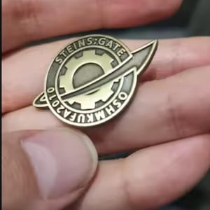 Peut inclure: Une broche circulaire en métal de couleur bronze avec un engrenage central entouré d'un anneau. Le texte STEINS;GATE est gravé en haut et OSHMKUFA2010 en bas. Un élément incurvé et pointu s'étend sur le côté droit de la broche.