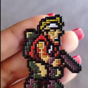 Peut inclure: Un pin's en émail au style pixel art représentant un personnage aux cheveux blonds, vêtu d'une chemise rouge et d'un pantalon sombre, tenant un objet sombre. Le design utilise une esthétique rétro avec des contours noirs et des couleurs vives.