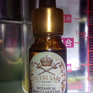 Puede incluir: Un pequeño frasco cuentagotas de vidrio ámbar con tapa dorada y una etiqueta que dice Putri Sari Herbs Botanical Skin Clarifying Infusion, con un gráfico de una corona y espadas cruzadas, frente a un recipiente medidor de plástico transparente.