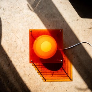 May include: A square lamp constructed from multiple stacked translucent orange acrylic sheets. The center features a circular, glowing yellow and orange light source. A white power cord extends from the side of the layered, geometric structure.