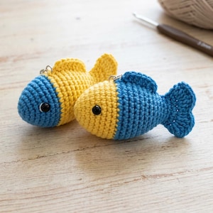 Puede incluir: Dos pequeños llaveros de pez tejidos a ganchillo a mano. Un pez tiene la cabeza azul y el cuerpo amarillo, mientras que el otro tiene la cabeza amarilla y el cuerpo azul. Ambos tienen ojos de cuenta negra y una argolla metálica en la aleta.