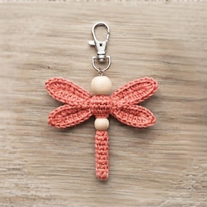 May include: A crochet dragonfly keychain featuring a coral-colored yarn body and four wings, accented with two smooth, light-colored wooden beads. The dragonfly is attached to a silver-toned metal lobster clasp for easy attachment to items.